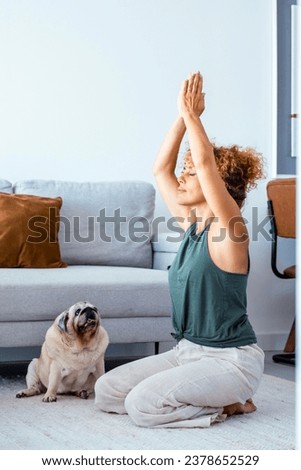 One healthy adult woman at home doing meditation exercise in yoga oriental zen position with her best friend dog pug sitting near her looking curious. Indoor leisure activity people in apartment