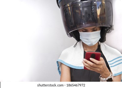 One Hispanic Young Adult Woman Sitting In A Hair Dryer Hood Beauty Salon Or Spa Using Protective Mask And Using Smartphone During Coronavirus Covid-19 Pandemic