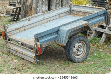 one iron open old industrial rusty gray dirty two-wheeled empty square car trailer stands on the ground near the cut tree trunks during the day on the street
