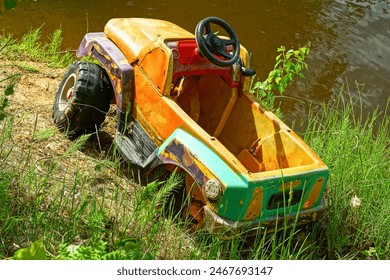 one large broken plastic yellow dirty discarded old children's car with two black wheels stands on the ground during the day on the street near the lake in the summer