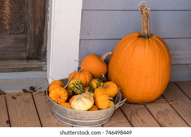 One large Pumpkin and Container of Smaller Pumpkins and Squash on the Porch of a Country Home
