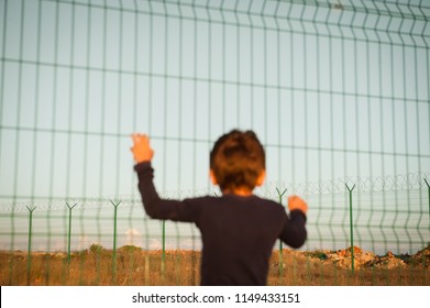 one little poor kid refugee holding high fence on border looking into far distance