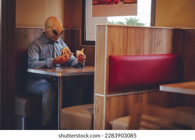 One man with alien mask eating alone inside a fast food store bad food hamburger and french fries. Ufo are among us living as human concept funny image. Invasion and diversity people indoor leisure