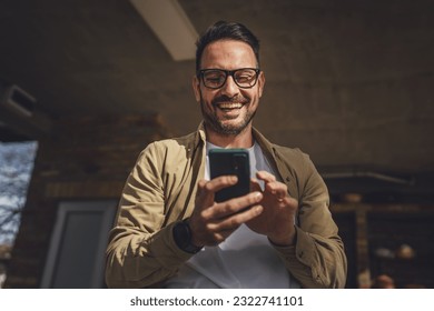 One man caucasian male with beard and eyeglasses sit outdoor in sunny day wear shirt use mobile smart phone for sms text messages browse internet online or video call use app copy space real people