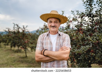 One man front view portrait of senior male farmer standing in the cherry orchard in summer day confident pensioner looking to the camera on his plantation wearing straw hat copy space - Powered by Shutterstock