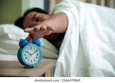 One man lying in bed extending his hand to snooze blue alarm clock on bedside table, typical morning routine
