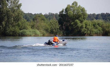 One man in orange lifejacket fast floats on inflatable motor boat with small outboard motor on the river on green trees background on far shore, active recreation on the water at Sunnny summer day