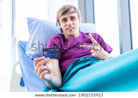 One a man patient on show holding a credit card lying with lifts two fingers up fighting on bed in the room hospital background,payment medical treatment concept