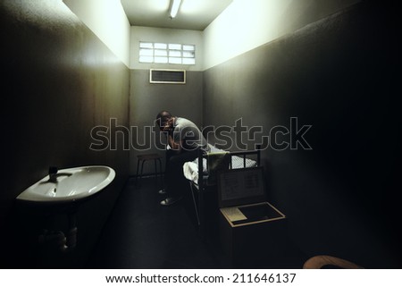 One man sitting on a bed in a small room of a dark prison.