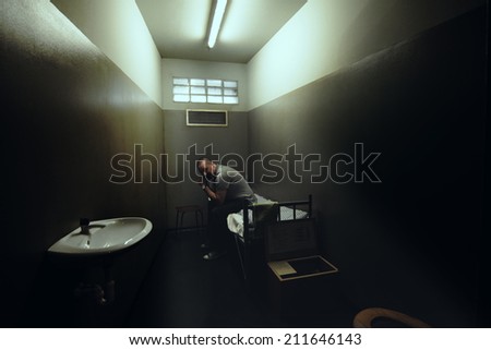 One man sitting on a bed in a small room of a dark prison.