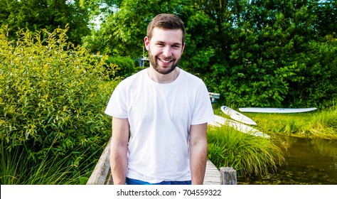 One Man In A White Tee Shirt And Blue Shorts Leaning On A Wooden Rail On A Pier, By The Grassy Edge Of A Lake. With Grass And Kayaks In The Background'