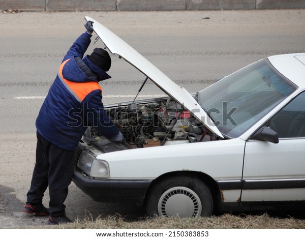 One man in work\
clothes repairs engine under hood of an old European car on the\
road. The problem with driving with the rise in price of spare\
parts in the economic\
crisis.