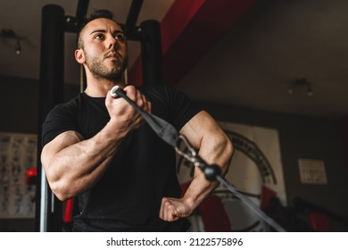 One man young adult caucasian male bodybuilder training arms bicep on the cable machine in the gym holding weight wearing black shirt dark photo real people copy space side view low angle