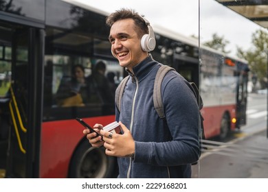 One man young adult male stand at public transport bus station taking bus with headphones and mobile smart phone in winter or autumn day with backpack student tourist city life copy space happy smile - Powered by Shutterstock