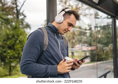 One man young adult male stand at public transport bus station waiting with headphones and mobile smart phone in winter or autumn day with backpack student or tourist city life copy space - Powered by Shutterstock