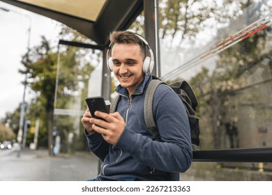 One man young adult male sit at public transport bus station waiting with headphones and mobile smart phone in winter or autumn day with backpack student or tourist city life copy space happy smile - Powered by Shutterstock