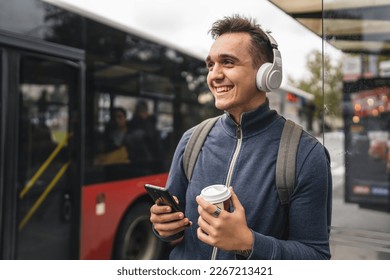 One man young adult male stand at public transport bus station taking bus with headphones and mobile smart phone in winter or autumn day with backpack student tourist city life copy space happy smile - Powered by Shutterstock