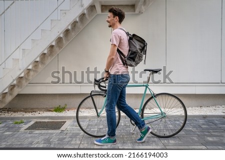 One man young adult male with brown hair and mustaches walking by the building with rucksack on his back and bicycle happy smile joyful real people copy space side view full length