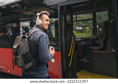One man young adult male stand at public transport bus station taking bus with headphones and mobile smart phone in winter or autumn day with backpack student or tourist city life copy space