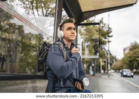 One man young adult male sit at public transport bus station waiting with headphones and mobile smart phone in winter or autumn day with backpack student or tourist city life copy space
