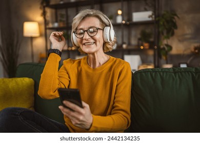 One mature blonde woman with headphones at home listen music online happy smile hold mobile phone smartphone use app application sit on the sofa bed - Powered by Shutterstock