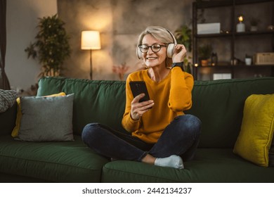 One mature blonde woman with headphones at home listen music online happy smile hold mobile phone smartphone use app application sit on the sofa bed - Powered by Shutterstock