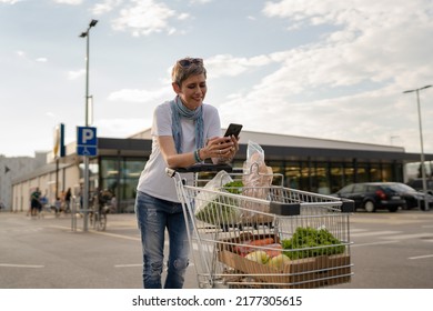 One Mature Woman Caucasian Female Walking In The Parking Lot In Front Of The Shopping Center Grocery Store Supermarket Chart Using Mobile Phone For Texting Or Checking The List Make A Call Copy Space