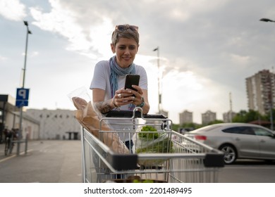 One Mature Woman Caucasian Female Walking In The Parking Lot In Front Of The Shopping Center Grocery Store Supermarket Chart Half Length Using Mobile Phone For Texting Or Checking The List Copy Space