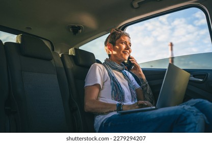 one mature woman caucasian female sitting on the back seat of the car working on laptop computer make a phone call talk in summer day with short gray hair modern on road wearing casual copy space - Powered by Shutterstock