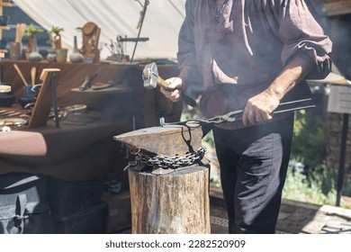 One middle-aged caucasian male blacksmith forges iron with a sledgehammer, standing behind a wooden stump on the street of Luxembourg city medieval castle, side view, close-up. The concept of medieval - Powered by Shutterstock