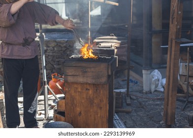 One middle-aged caucasian male blacksmith melts iron in fire holding it with tongs, standing behind a wooden stand on a Luxembourg city street in a medieval castle side view, close-up. The concept of  - Powered by Shutterstock