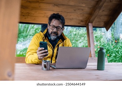 One modern mature freelance using phone and laptop outdoor in forest cabin enjoying nature and freedom in business travel lifestyle. Adult man messaging outside. Wearing yellow jacket and glasses - Powered by Shutterstock