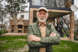 One Modern Senior Caucasian Man Portrait Of Male Grandfather With Beard And Hat Cap Stand In Front Of His House In Day Wear Jacket Happy Confident Copy Space Arms Crossed