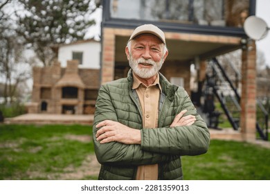 One modern senior caucasian man portrait of male grandfather with beard and hat cap stand in front of his house in day wear jacket happy confident copy space arms crossed
