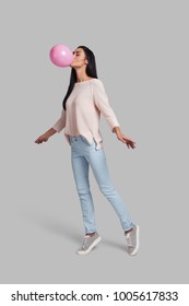One more step and she will fly. Full length studio shot of attractive young woman in casual wear blowing up a pink balloon while standing against grey background