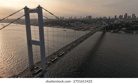 One of Turkey’s most recognizable transportation structures, the 15 July Martyrs Bridge stretches diagonally across the Bosphorus in this wide-angle drone image. Formerly known as the Bosphorus Bridge, it forms a scene of high architectural and scenic value together with the bright sunlight reflected on the water. Dense urban texture appears on one side of the bridge while open water fills the other, and vehicle traffic can be seen moving in a steady flow. Boats and small ships moving on the sea make marine traffic visible and add a second layer of motion to the composition. This frame is especially powerful because it simultaneously tells the geography of Istanbul, the connection between two continents, the shoreline and modern urban life. With near-daylight golden tones and a soft sky, the scene feels both calm and iconic as an Istanbul panorama.