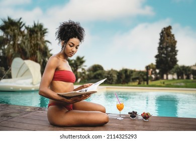 One multiracial beautiful girl with curly hair in red biking relax 
ing sitting poolside reading a magazine and having a healthy fruit breakfast cherries and strawberries - copy space 