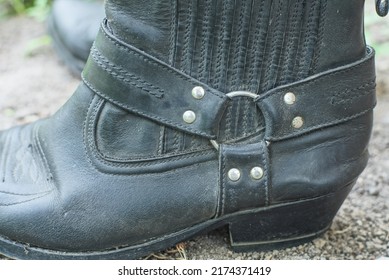 one old black worn fashionable leather cowboy boot with leather buckles and metal studs stands outside during the day