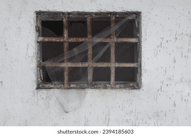 one old gray window behind rust eaten iron grating on a white concrete wall. old and aged window frame. plastic net. exterior. 