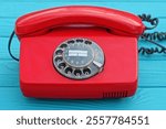 one old plastic red with round disk and numbers and handset with speakers analog with black cord office classic telephone stands on wooden green table