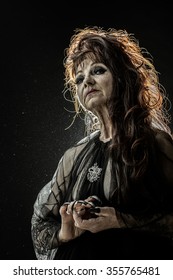 One Old Scary Smiling Witch Woman With Long Red Knotty Hair In Blouse With Black Long Nails On Hand As Halloween Character With Bright Makeup In Studio On Black Background, Vertical Photo