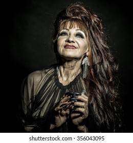 One Old Scary Smiling Witch Woman With Long Red Knotty Hair In Blouse With Black Long Nails On Hand As Halloween Character With Bright Makeup In Studio On Black Background, Square Photo