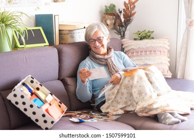 One Old Woman With Gray Hair, Remembers The Past Looking At Old Photographs Of A Lifetime. Old Mold Storage. Sitting On The Sofa With A Mobile Phone And Tablet.