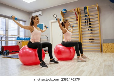 One on one training, young woman with her fitness trainer sitting on swiss ball and holding weights 