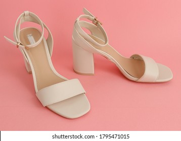 One pair of white women's sandals with high and square heels stands on a pink paper background, close-up side view.