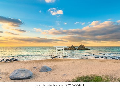 One person praying on the Itoshima Beach of Fukuoka standing in front of a white Shinto torii gate leading to the famous Sakurai Futamigaura's Meoto Iwa Couple Rocks in the evening sunset light. - Shutterstock ID 2227656431