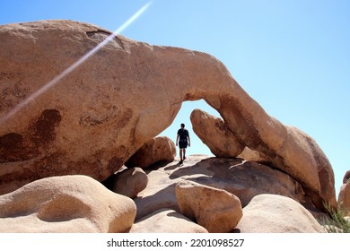 One person walking arch rock inside Joshua Tree national park. Man hiking under impressive stone and rock formation with blue skies in background. Adventurer in south California during hot summer.