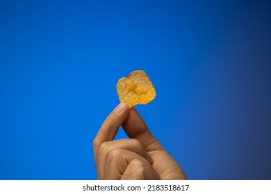 One Potato Chip Held In Hand By Caucasian Male Hand. Close Up Studio Shot, Isolated On Blue Background.