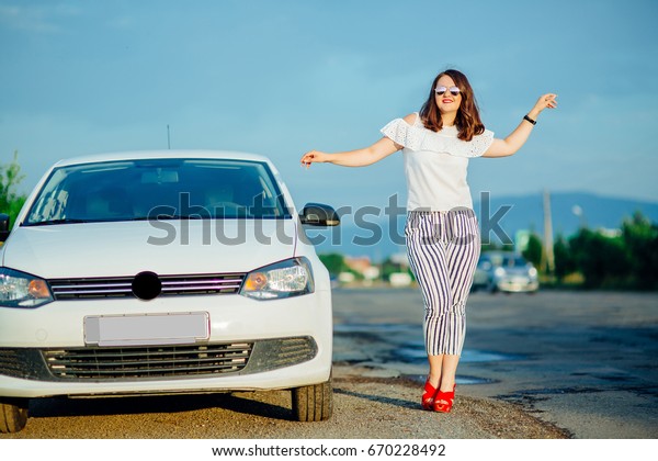 One pretty\
sensual sexy thinking straight slim fashionable young woman with\
long legs in red high heeled shoes stands on road near beautiful\
car white color outdoor sunny day,\
