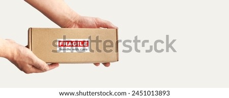 One rectangular cardboard box in hands for fragile items packing on a plain light background. An adhesive red warning label, sticker for packaging 'Fragile, Handle with care, Thank you'. No people